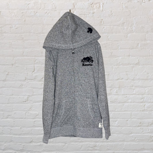 Grey Roots full-zip hooded sweatshirt with logo on chest and maple leaf on hood, hanging against a white brick wall.