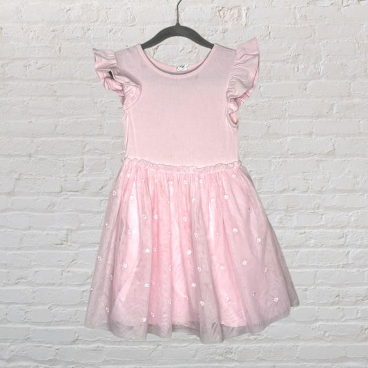 Pink dress with ruffle sleeves and a tulle skirt, featuring embroidered floral details, hanging against a white brick background.