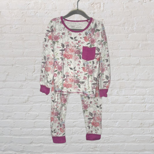Women's floral print pajama set with pink cuffs and pocket, hanging on a hanger against a white brick background.