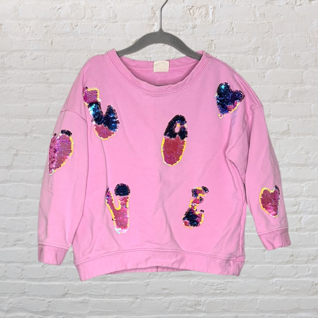 Pink sweatshirt featuring colorful sequin heart and letter designs, displayed on a hanger.