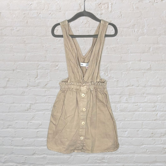 Beige button-front pinafore dress with adjustable straps on a hanger, displayed against a brick wall.