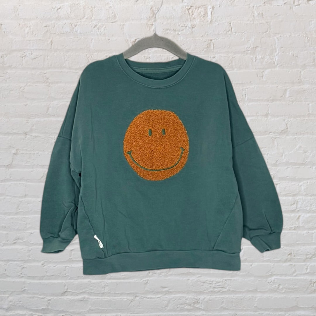 Teal sweatshirt with an orange textured smiley face design on the front, hung on a hanger against a white brick wall.