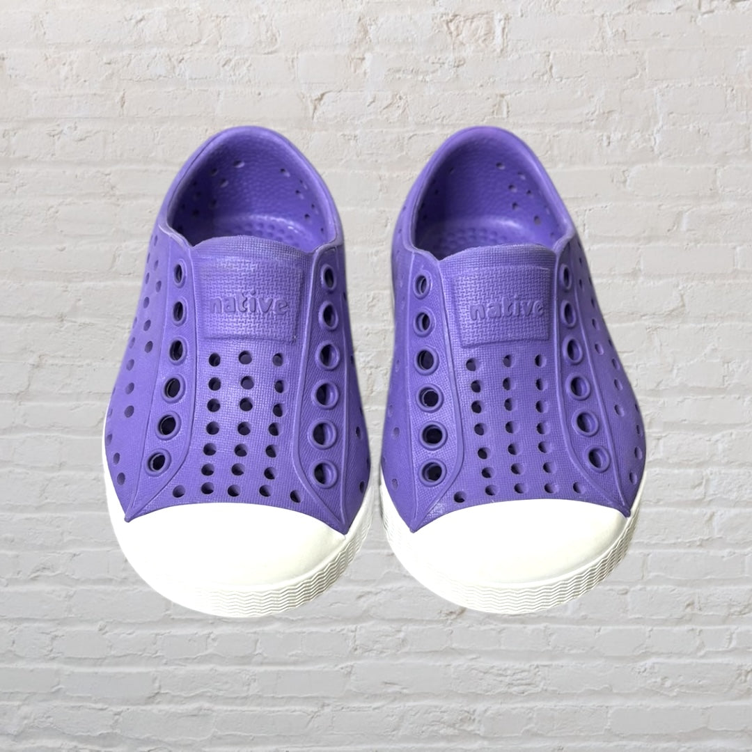 Purple perforated slip-on shoes with white sole, designed by Native, against a white brick background.