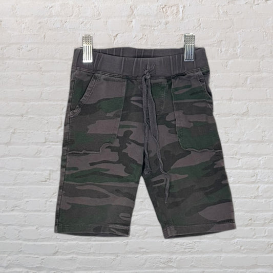 Camouflage print shorts with a drawstring waistband, displayed on a hanger against a white brick wall.