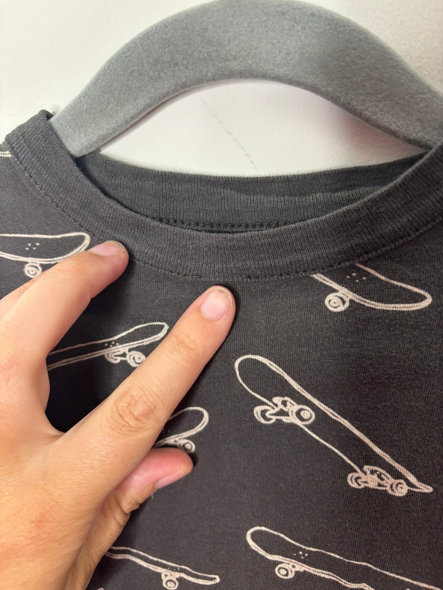 Black T-shirt with all-over white skateboard print hanging on a hanger against a white brick wall.