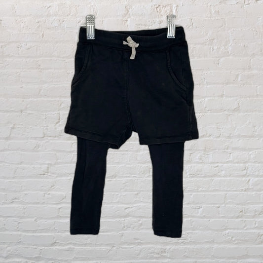 Children's black athletic shorts with attached leggings and a drawstring waist, hanging against a brick wall.