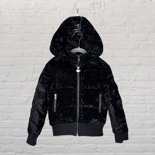 Black velvet hooded jacket with zipper front and two side pockets, displayed on a hanger against a brick wall.