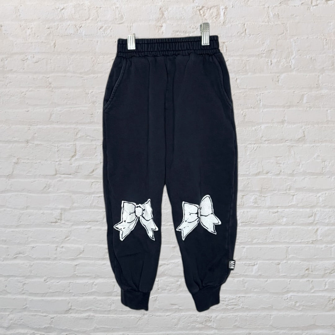 Black sweatshirt and pants set featuring large white bow graphics, displayed on a hanger against a brick wall.