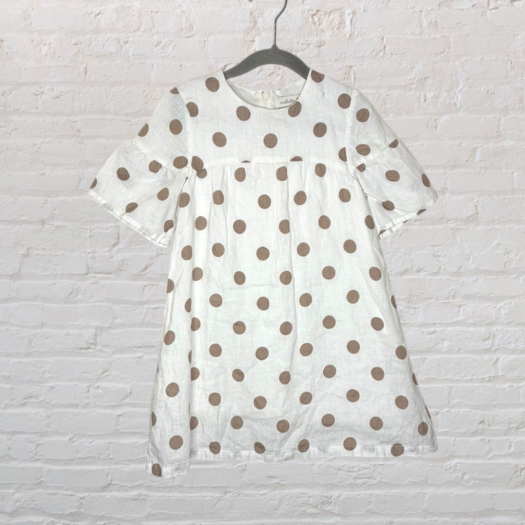 White dress with brown polka dots and short sleeves hanging on a black hanger against a white brick wall.
