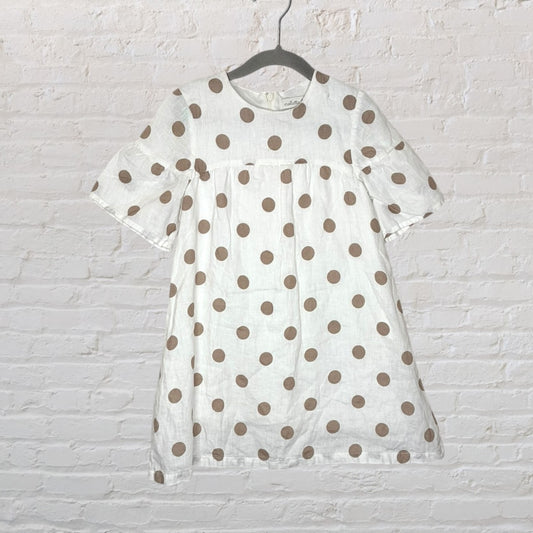 White dress with brown polka dots and short sleeves hanging on a black hanger against a white brick wall.