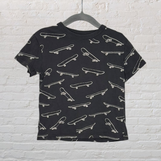 Black T-shirt with all-over white skateboard print hanging on a hanger against a white brick wall.