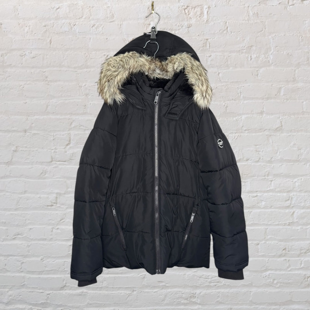 Black puffer jacket with a faux fur hood, hanging against a white brick wall.