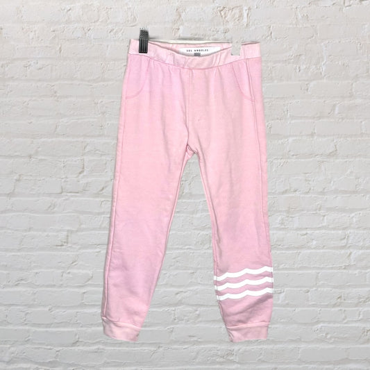 Light pink jogger sweatpants from Sol Angeles with three white wave stripes on one leg, displayed against a brick wall.