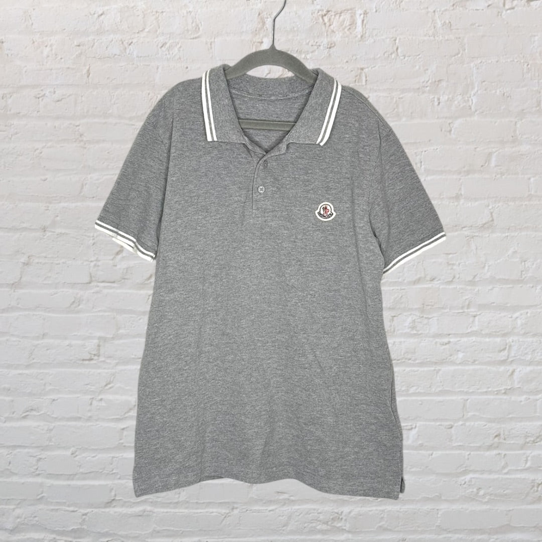 Grey polo shirt with a Moncler logo patch on the chest, featuring white striped collar and sleeve cuffs, hanging on a hanger.