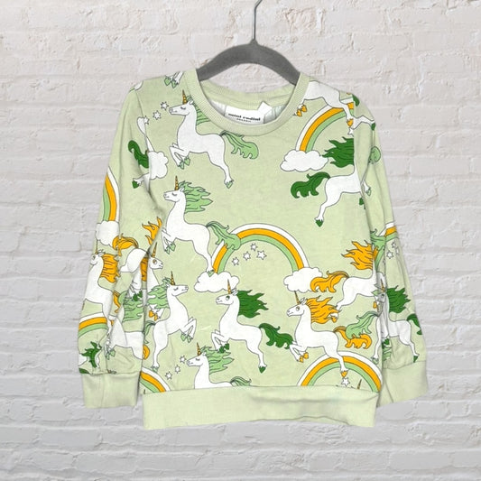 A light green sweatshirt with a playful unicorn and rainbow print pattern, hung on a black hanger against a brick wall.