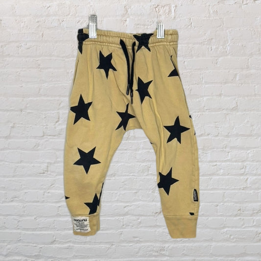 Beige jogger pants with black star prints, elastic waistband, and cuffs, hanging against a brick wall.