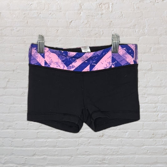 Black athletic shorts with an abstract pink and purple patterned waistband, displayed against a white brick background.