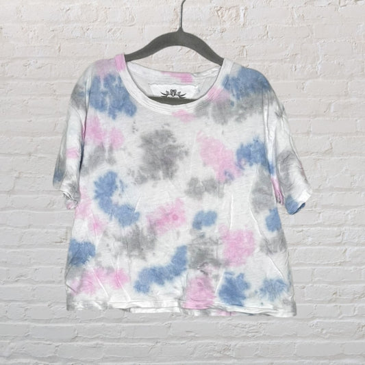 Unisex tie-dye T-shirt with blue, pink, and gray patterns, displayed on a hanger against a brick wall.