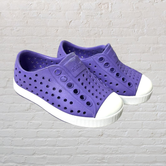 Purple perforated slip-on shoes with white sole, designed by Native, against a white brick background.