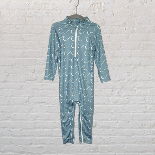 Blue onepiece swimsuit with white wave patterns and a front zipper, displayed on a hanger against a white brick wall.