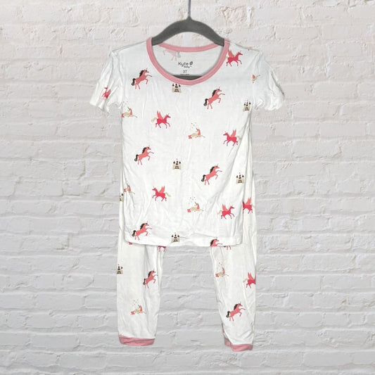 White pajama set with pink unicorn prints, short sleeves and pants, by Kyte, hanging against a brick wall.