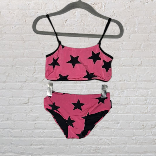 Pink bikini set with black star pattern hanging on a hanger against a white brick wall.