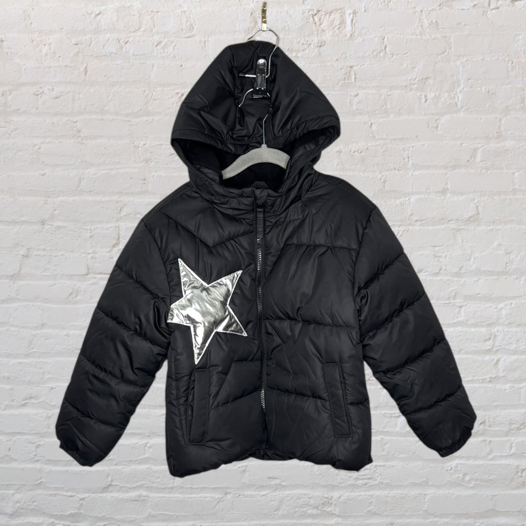Black puffer jacket with hood and a large metallic silver star design on the front, hanging against a brick wall.