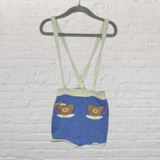 Knit suspender shorts in blue with green trim, featuring embroidered tea cups, hung on a hanger against a brick wall.