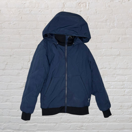 Navy blue hooded puffer jacket with black cuffs and hem, displayed on a hanger against a white brick wall.