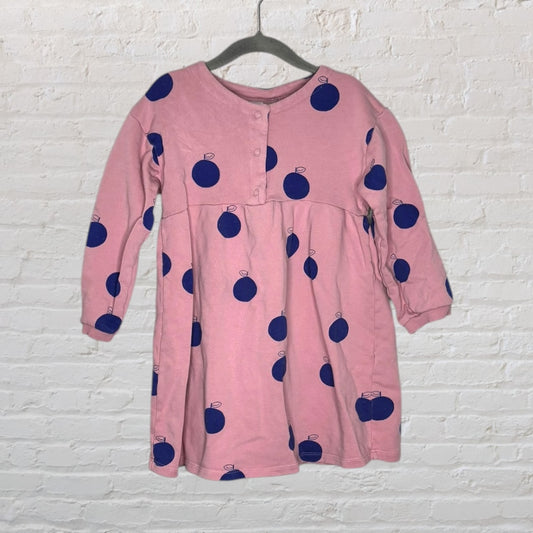 Pink long-sleeve dress with blue apple print, hanging on a hanger against a white brick wall.