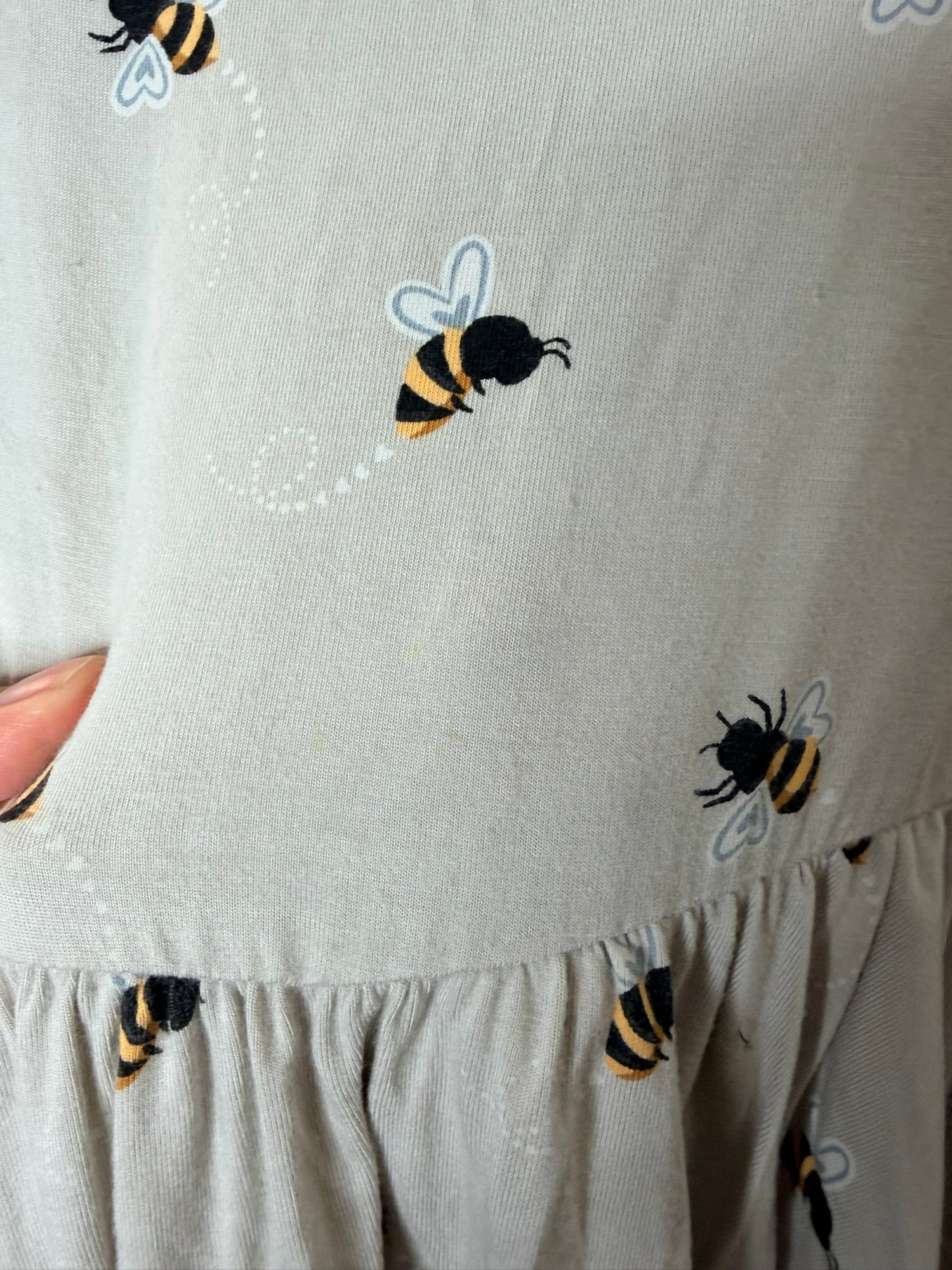 Light grey skater dress with bee patterns, hanging on a hanger against a white brick wall.