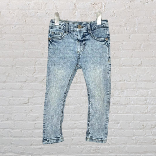 Light wash denim jeans for kids, featuring classic pocket design and button closure, hung against a white brick wall.