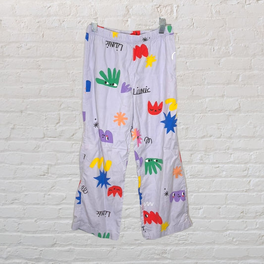 Lavender pajama pants with colorful abstract patterns and playful graphics, hanging against a white brick wall.