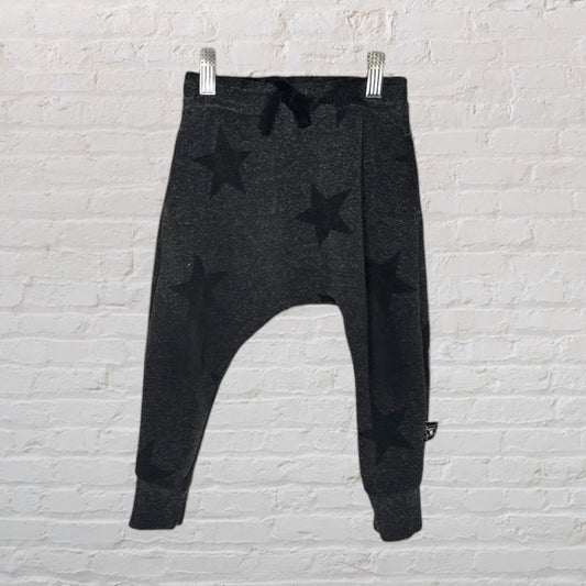 Dark gray jogger pants with black star patterns, hanging on a white brick wall.