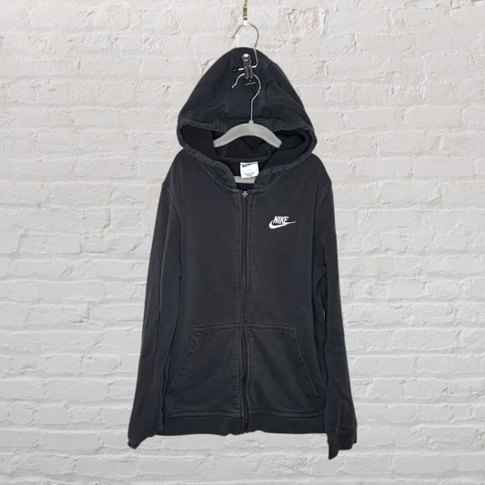 Black Nike zip-up hoodie with a white logo on the chest, hanging on a hanger against a brick wall.