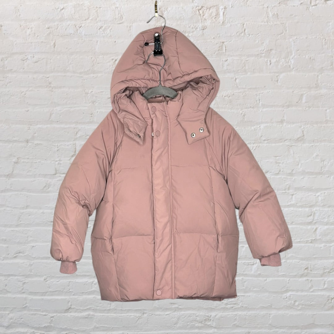 Pink hooded puffer jacket with front snap buttons, displayed on a hanger against a brick wall.