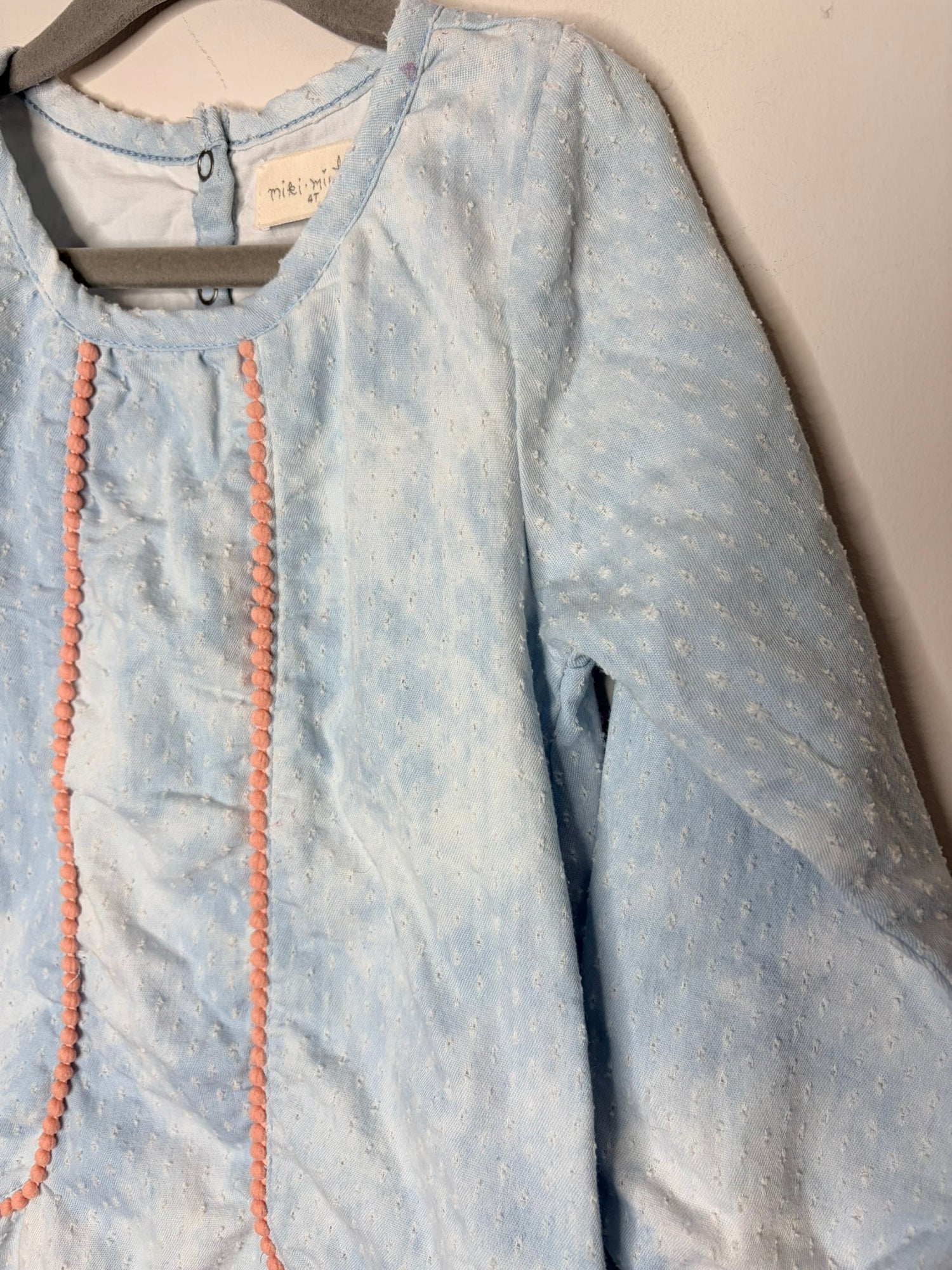Light blue long sleeve dress for kids with ruffle hem and pink trim, hanging on a gray hanger.