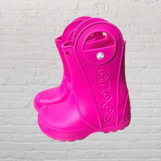 Bright pink Crocs rain boots with handles, featuring the Crocs logo on the side against a white brick background.