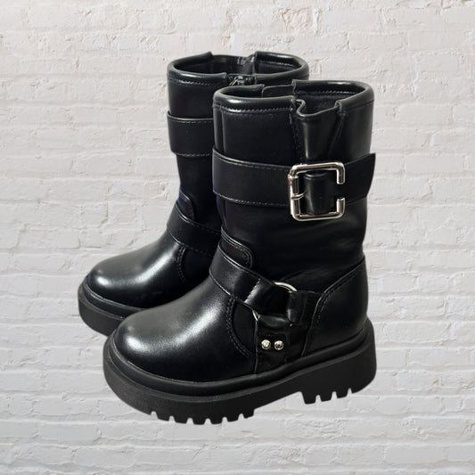 Pair of chunky black boots with silver buckle accents and a thick sole against a white brick background.