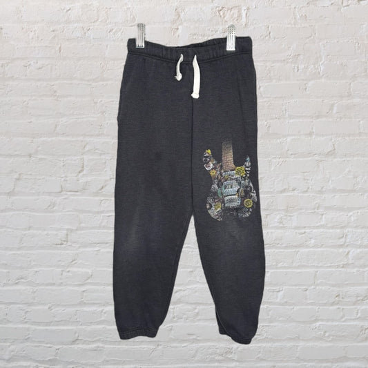 Dark grey sweatpants featuring a colorful graphic of a guitar on the leg, with a drawstring waistband, hanging against a light brick wall.