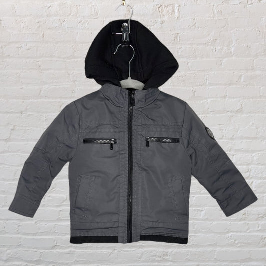 A children's gray hooded jacket with front zippers and black hood, displayed on a hanger with a white brick background.