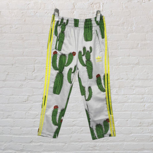 Adidas track pants with cactus print, featuring yellow stripes on the sides, against a white brick background.