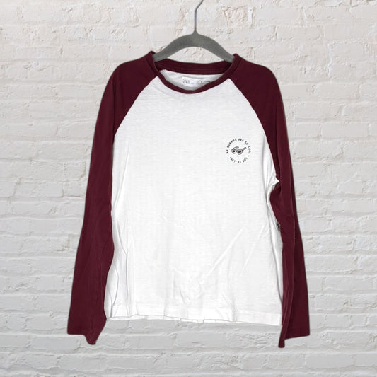 White and burgundy long-sleeve raglan t-shirt with a small graphic print on the left chest, hanging on a gray hanger.