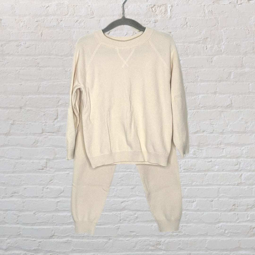Cream-colored knit sweater and pants set hanging on a hanger against a white brick wall.