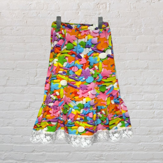 A vibrant multicolor skirt with a candy print design and white lace hem, hanging against a white brick wall.