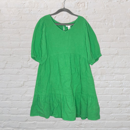Green V-neck dress with puff sleeves and tiered design, hanging against a white brick background.