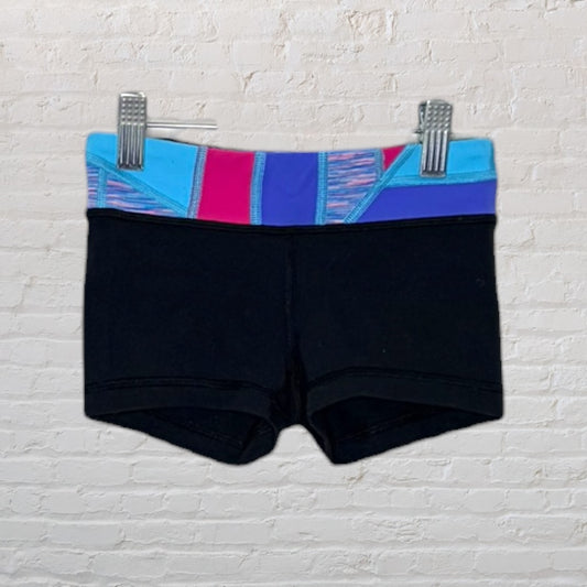 Black athletic shorts with a colorful blue, purple, and pink waistband, clipped on display against a white brick wall.