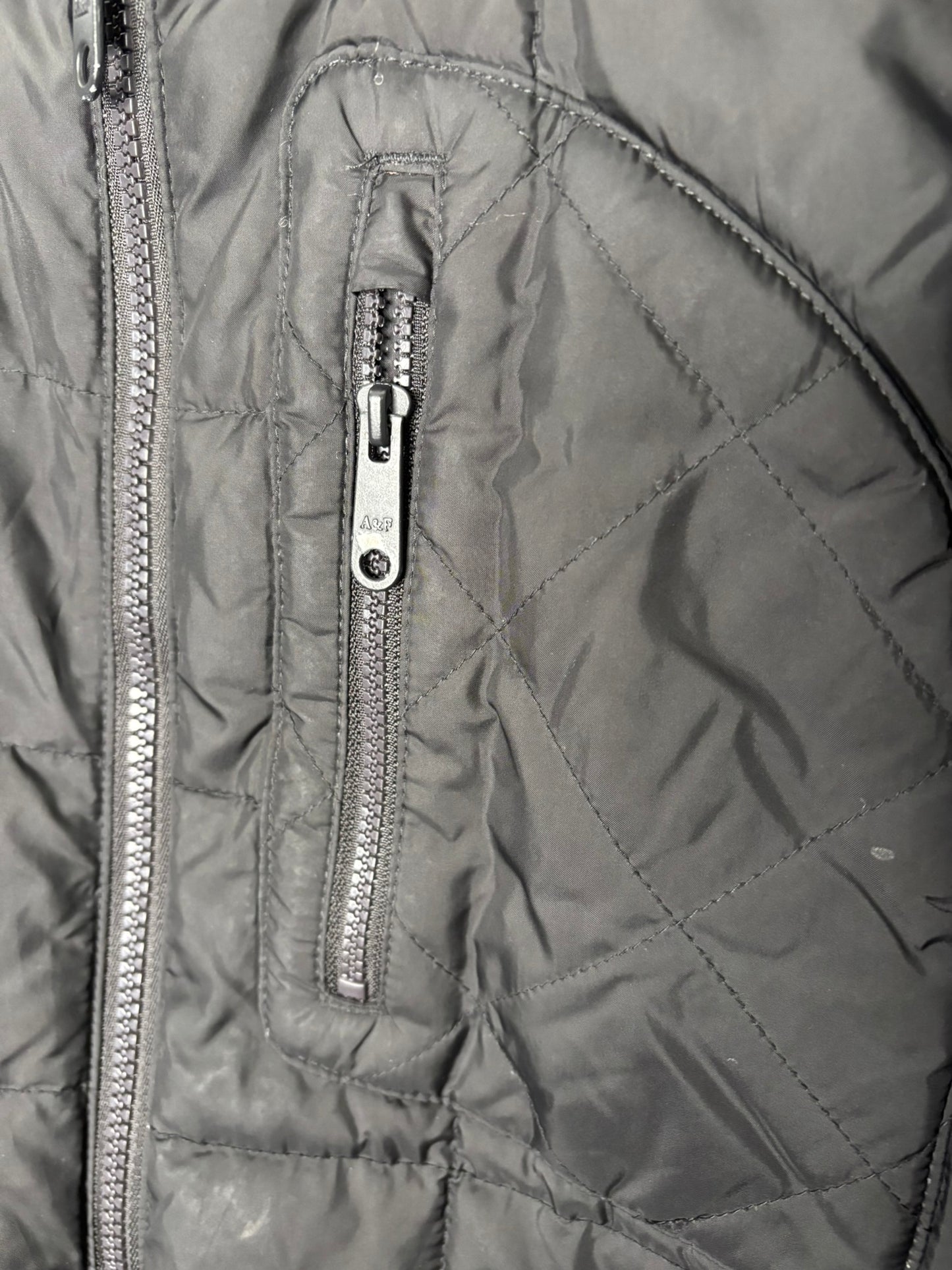 Black quilted puffer jacket with zipper pockets and a round patch on the sleeve, hanging against a white brick wall.