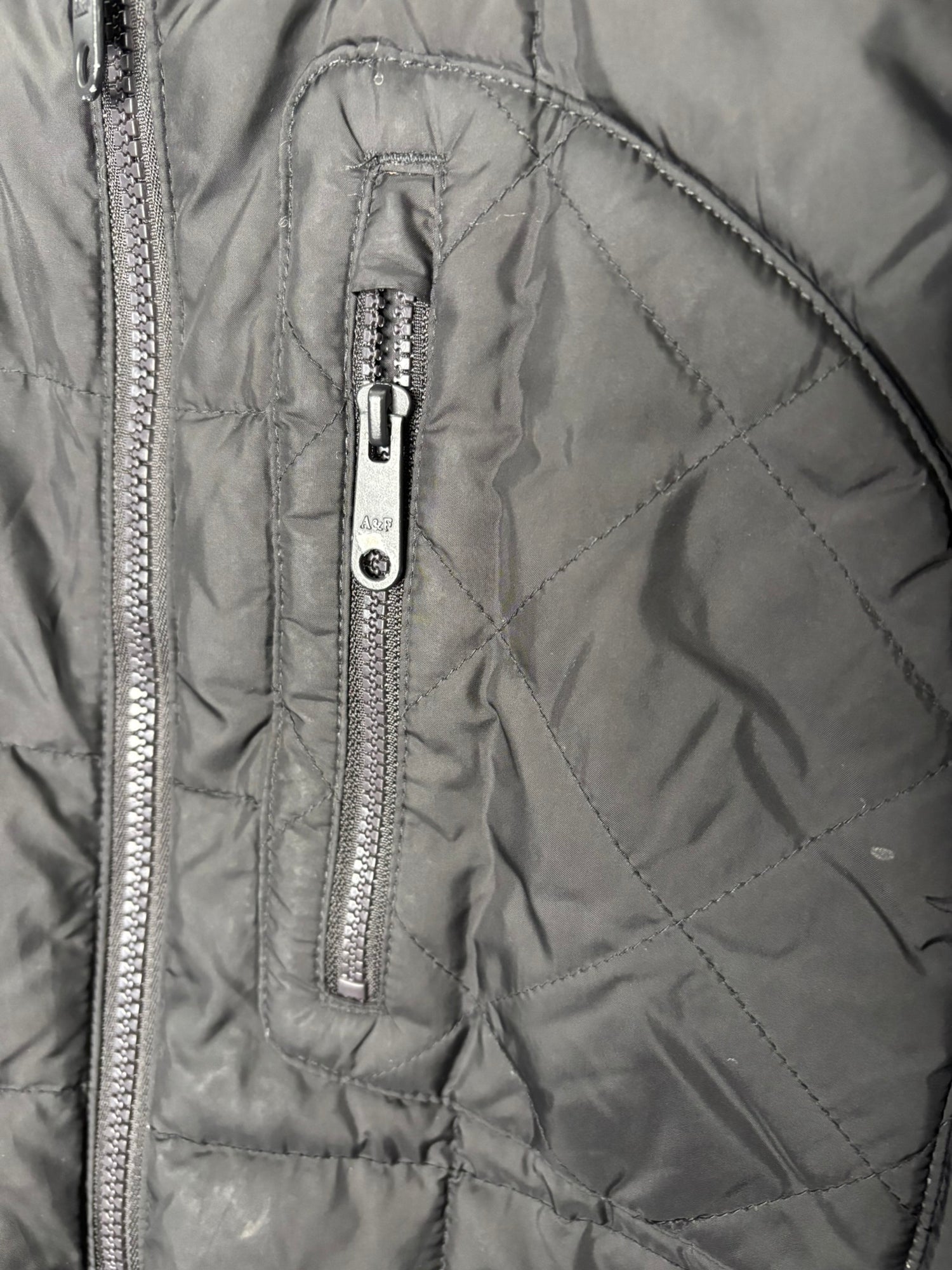Black quilted puffer jacket with zipper pockets and a round patch on the sleeve, hanging against a white brick wall.
