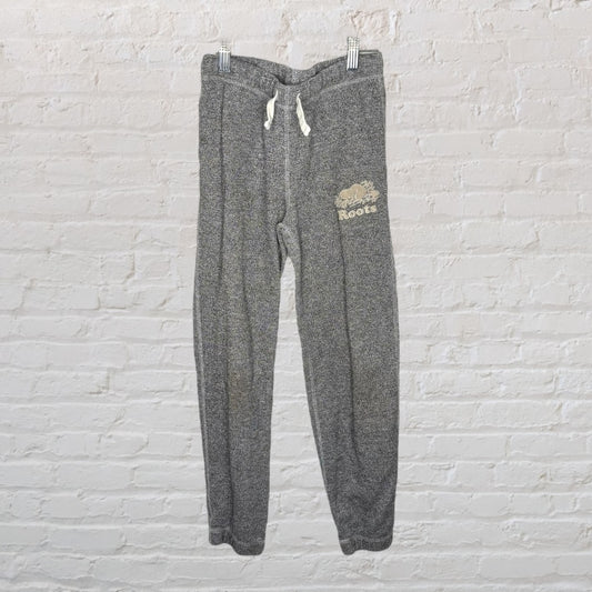 Heather grey Roots sweatpants with logo, white drawstrings, hung against a white brick wall.